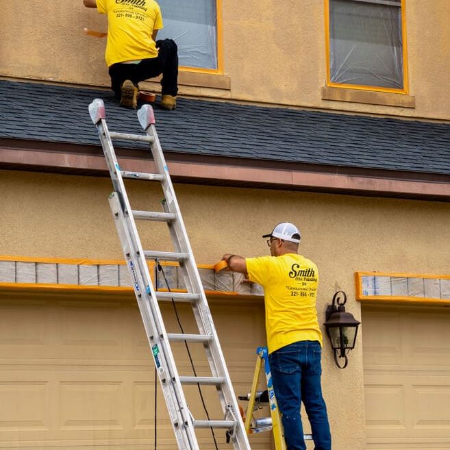 Two Smith and Co. painters in yellow shirts prepping and painting a home exterior